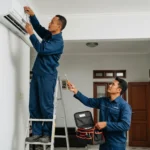 Dua teknisi AC bekerja sama memperbaiki unit AC.